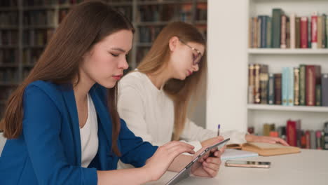 una mujer navega por internet en una tableta en el vestíbulo de la biblioteca. una mujer busca información en internet cerca de una amiga escribiendo la tarea en la mesa. educación para estudiantes