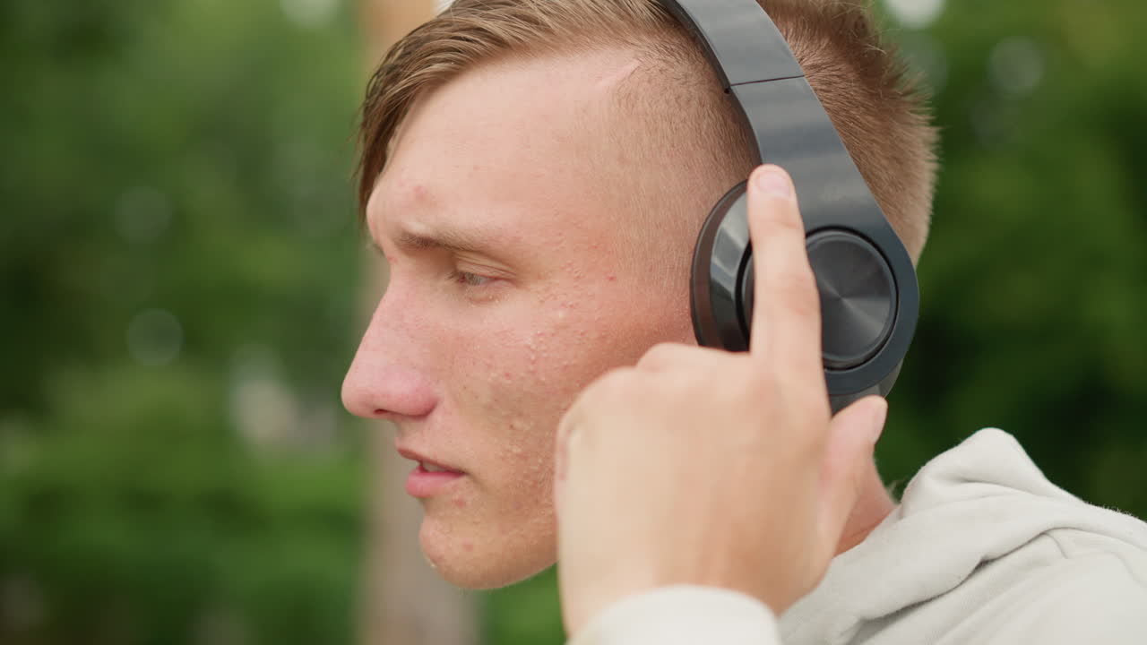 un hombre blanco joven con auriculares en un parque lluvioso, con el pelo y la sudadera con capucha mojados, ajustando los auriculares y tocando los controles mientras escucha música con atención; un primer plano muestra un peinado undercut con gotas de lluvia