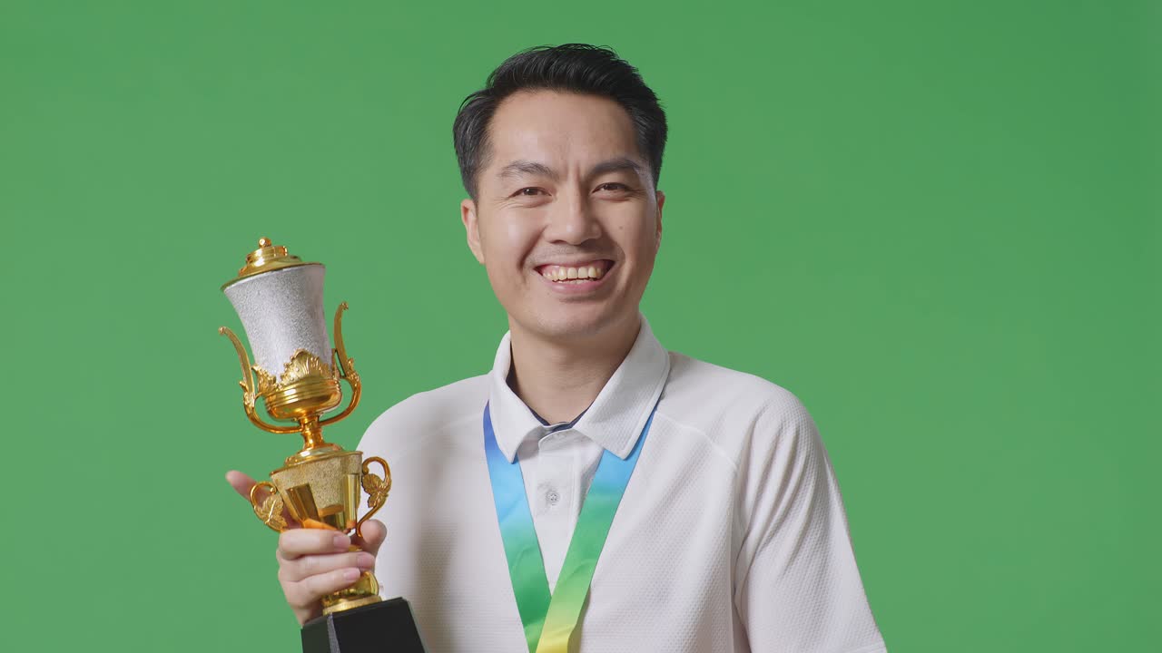 가까이에서 아시아 남자가 금메달을 입고 손에 금 트로피를 들고 첫 번째 우승자로 스튜디오의 녹색 화면 배경에서 카메라에 미소 짓고 있습니다.