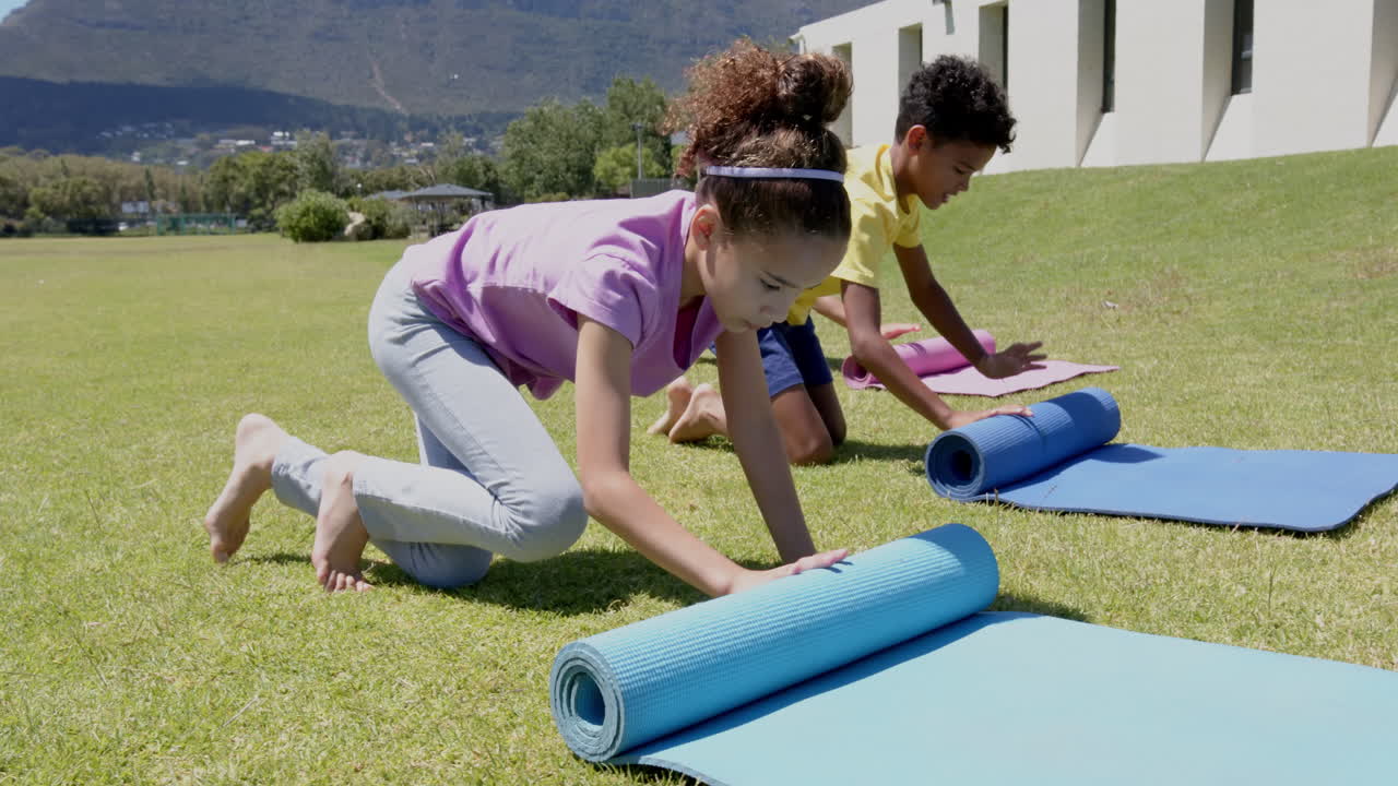 Biracial girl in a purple shirt is rolling up a blue yoga mat outdoors in school
