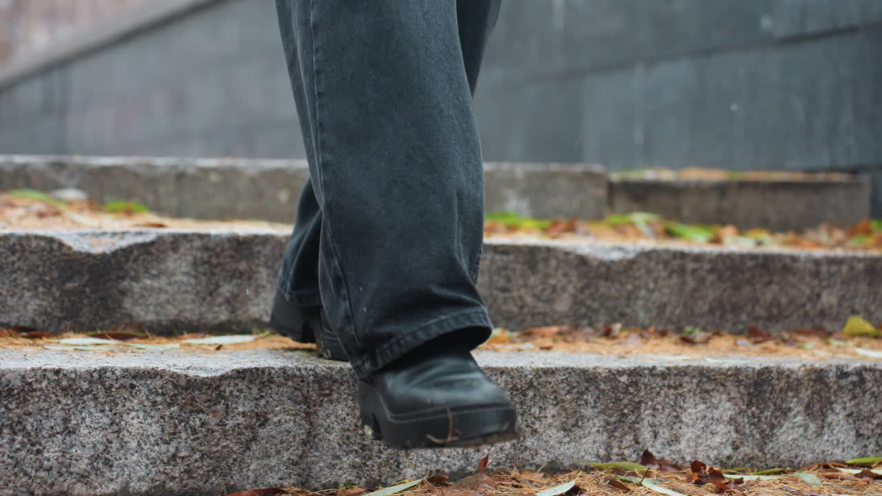 Legs of person walking down stone steps during light snowfall, wearing black jeans and black boots, with colorful autumn leaves scattered along the path creating a seasonal atmosphere