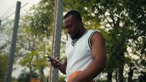 A-guy-with-black-skin-in-a-white-T-shirt-smiles,-holds-a-basketball-in-his-hand-and-types-a-message-on-a-smartphone