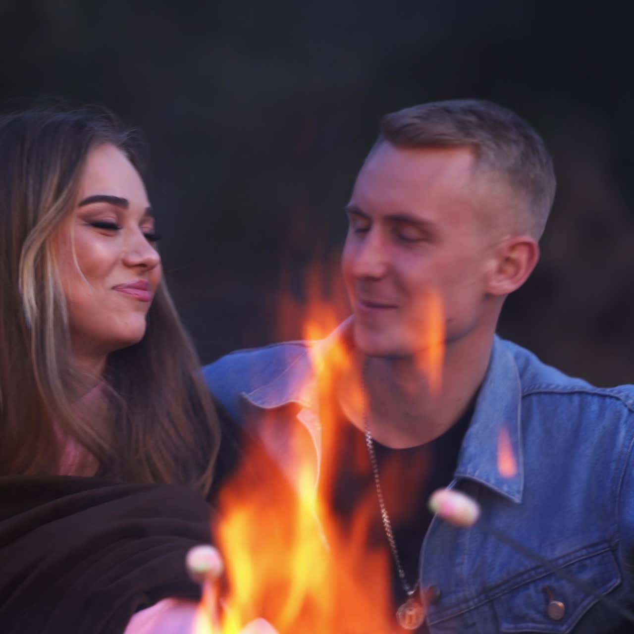 Happy young couple sitting and hugging near the campfire and talking. Dating pair holding barbecued marshmallows