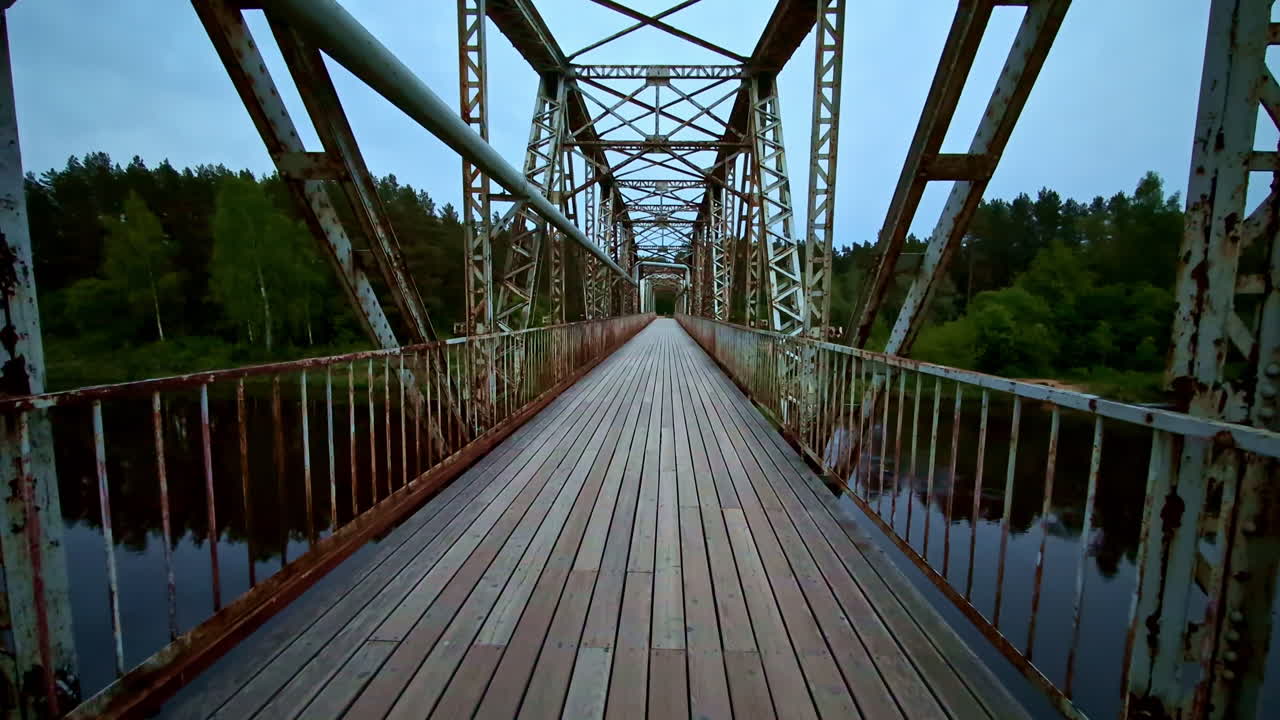Rusty steel bridge with wooden planks crossing river surrounded by trees at dusk