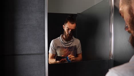 joven barbudo mirando al espejo y aplicando máscara de crema limpiadora negra en la cara en el baño