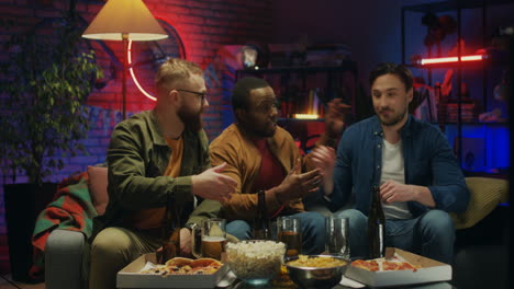 young handsome man coming to his multiethnic friends in the dark living room, drinking beer with snacks and watching match on tv sport channel.