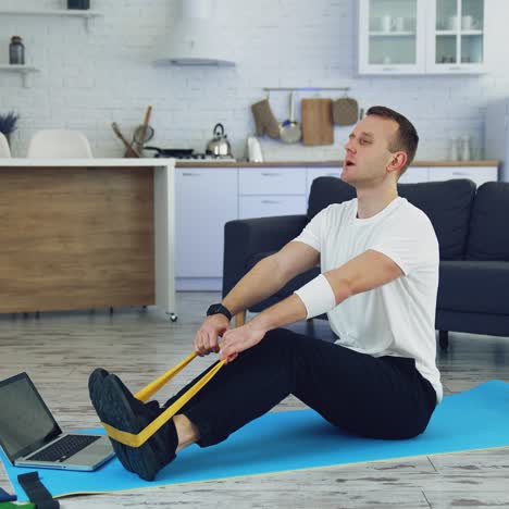 workout at home during quarantine. young man is doing breath exercises with fitness rubber band on the floor in front of laptop. sporty lifestyle indoors