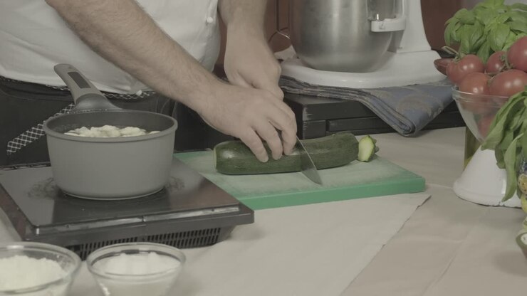 Primo piano delle mani dello chef che tagliano zucchine verdi con un coltello accanto a una pentola di acqua bollente.