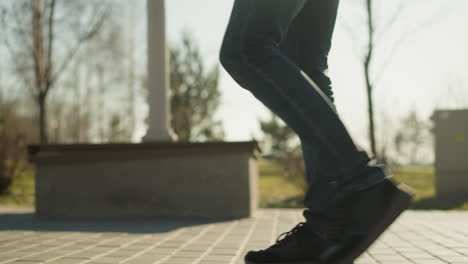 a close-up leg view of an adult walking close to an open field with trees, on a paved path, wearing jeans and black sneakers