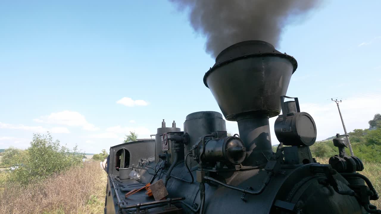 A historic steam engine is chugging through the rural countryside on a sunny day. The powerful locomotive travels on the railway, billowing dark smoke, starting an exciting journey