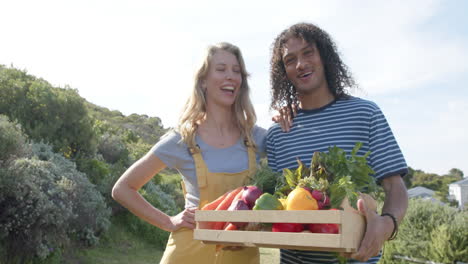 portrait of happy diverse couple holding basket of fresh vegetables in garden, slow motion