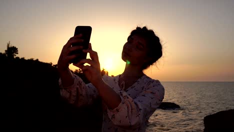 adorable caucasian brunette girl in light dress make selfie at the cliff above the sea at sunrise time