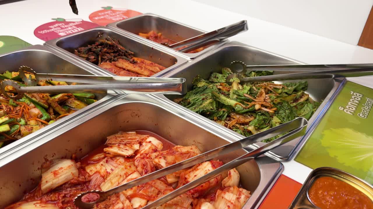 Hand uses tongs to pick kimchi and side dishes at a brightly lit buffet counter
