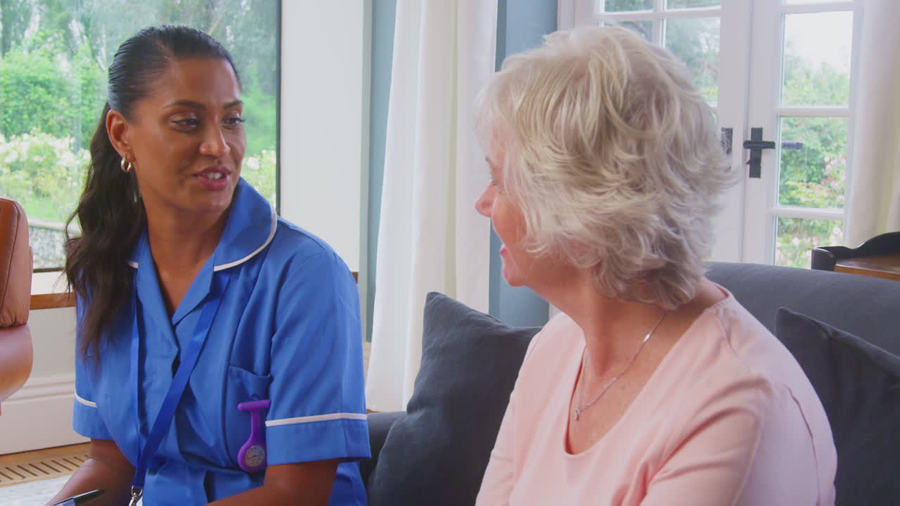 Senior Woman At Home Talking To Female Nurse Or Care Worker In Uniform Making Notes On Clipboard