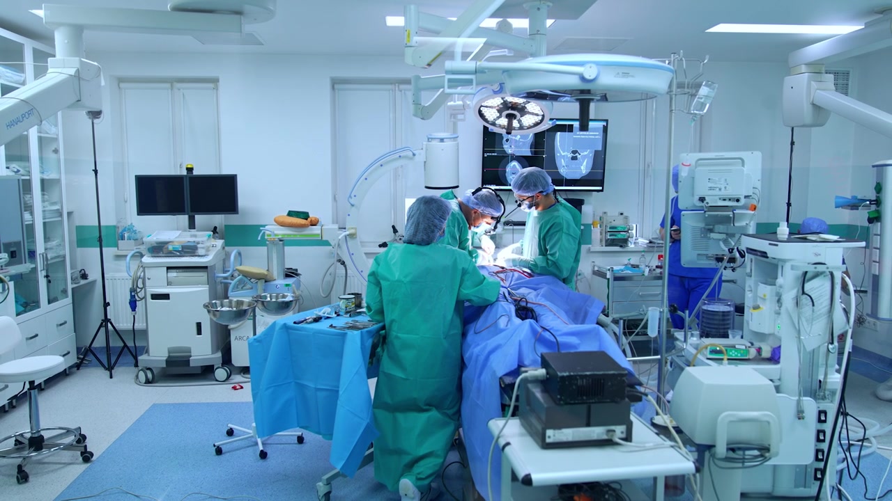 Spacious surgery room with advanced medical equipment. Two surgeons and female scrub nurse perform operation at the operating table.