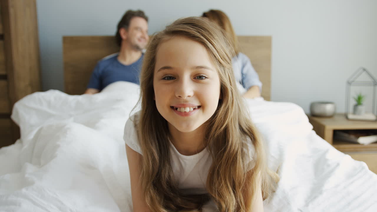 귀여운 어린 소녀가 누워 카메라를 보고 웃고 있고, 그녀의 부모는 그녀 뒤에 있는 침대의 담요 아래에 누워 있다