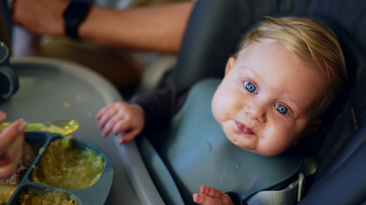 Adorable baby boy with a bib sits at the table. Unrecognized mom gives pureed food to a child. Top view.