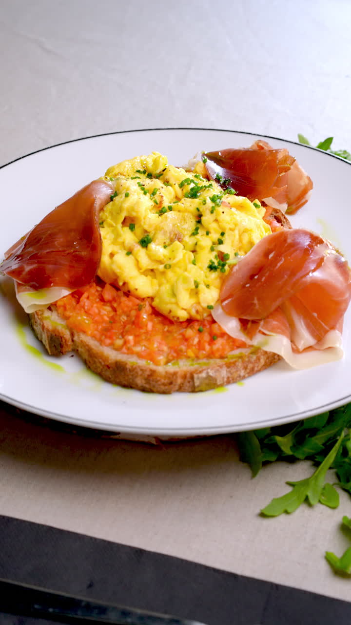Eastern European cuisine presents scrambled eggs with prosciutto and vegetable spread, served on toasted bread, garnished with herbs, accompanied by a fork and knife on a set table
