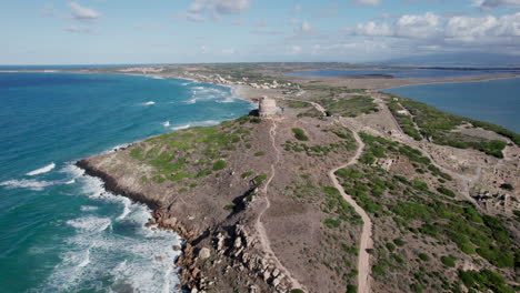 Torre-di-San-Giovanni-di-Sinis,-Sardinia:-aerial-view-travelling-out-over-the-famous-tower-and-overlooking-a-spectacular-coastline-with-turquoise-waters