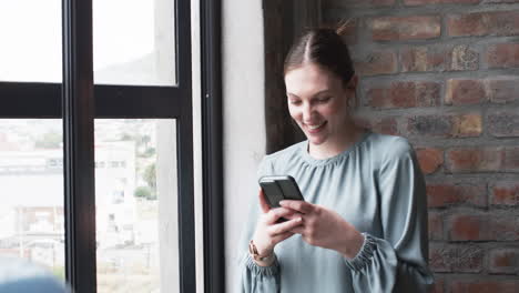 young caucasian businesswoman smiles while using her smartphone by a window