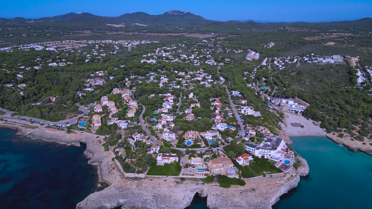 Stunning drone view of a luxury residential real estate area on the coast of Mallorca, Spain. The scene shows villas, resort hotels, and homes built on the cliffside overlooking the Mediterranean Sea
