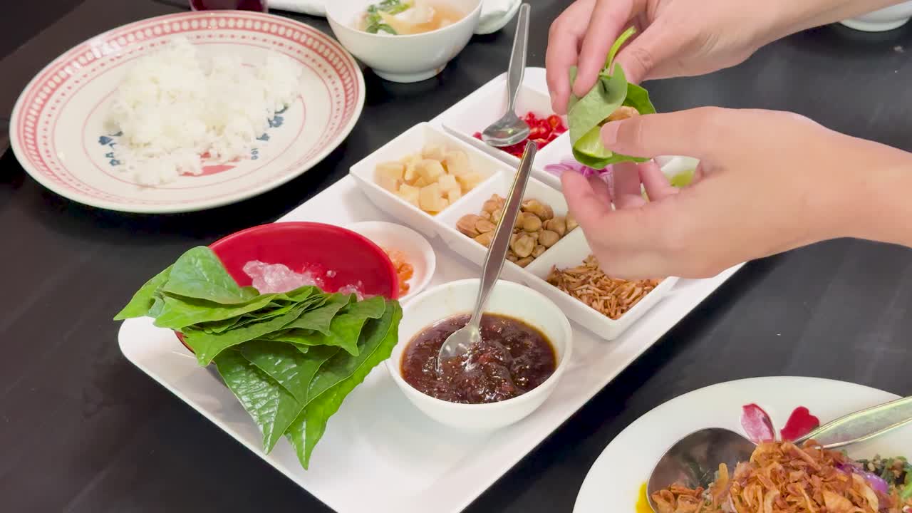 Hands prepare traditional Miang Kham by adding ingredients to wild betel leaves on a dining table, under bright, even lighting with a steady overhead camera view