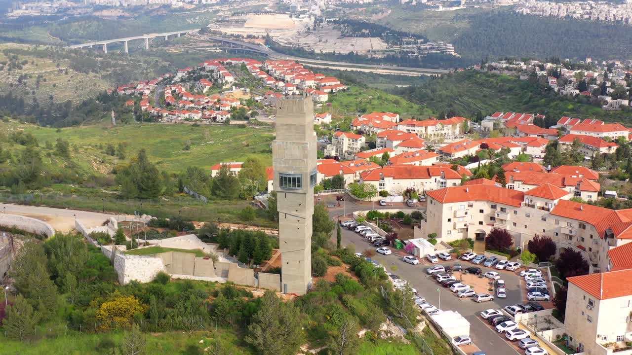 Aerial view of a residential neighborhood featuring a tall concrete tower amidst red-roofed houses and green hills