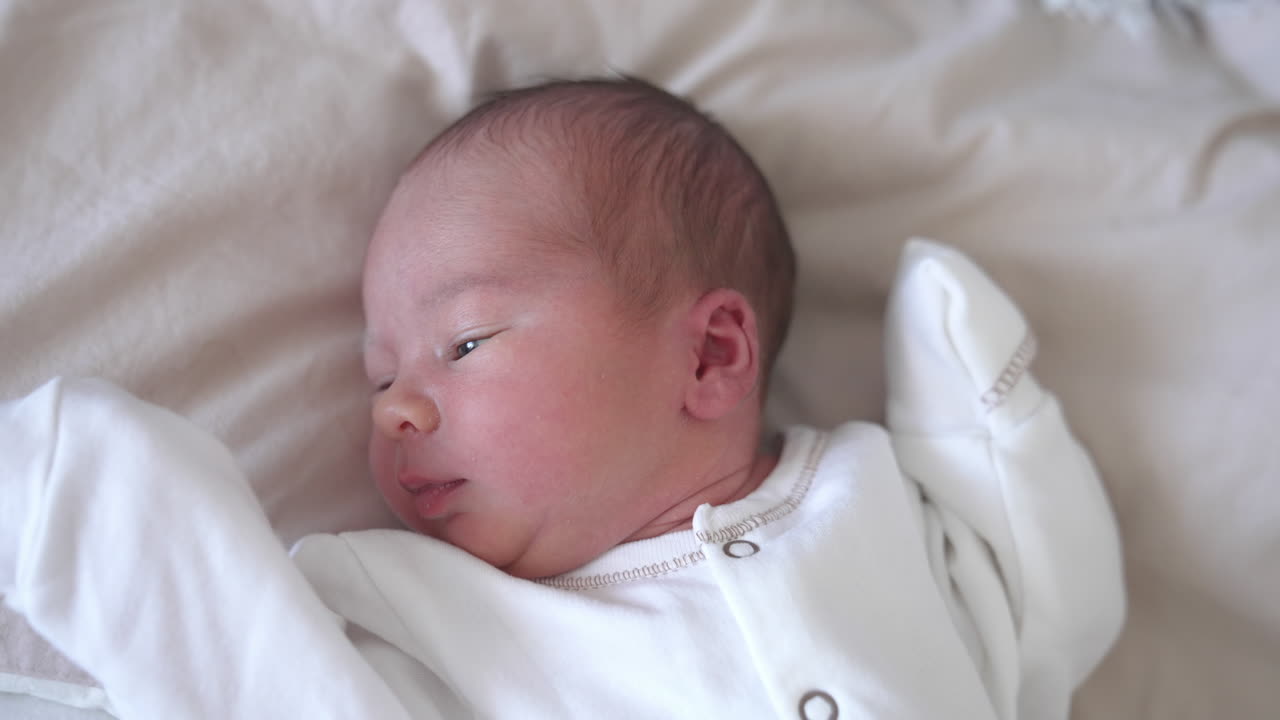 Sweet newborn boy is lying on the bed and turning his little head back and forth. Yawning tiny kid in a white costume. Close-up from above.