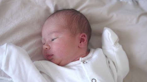sweet newborn boy is lying on the bed and turning his little head back and forth. yawning tiny kid in a white costume. close-up from above.