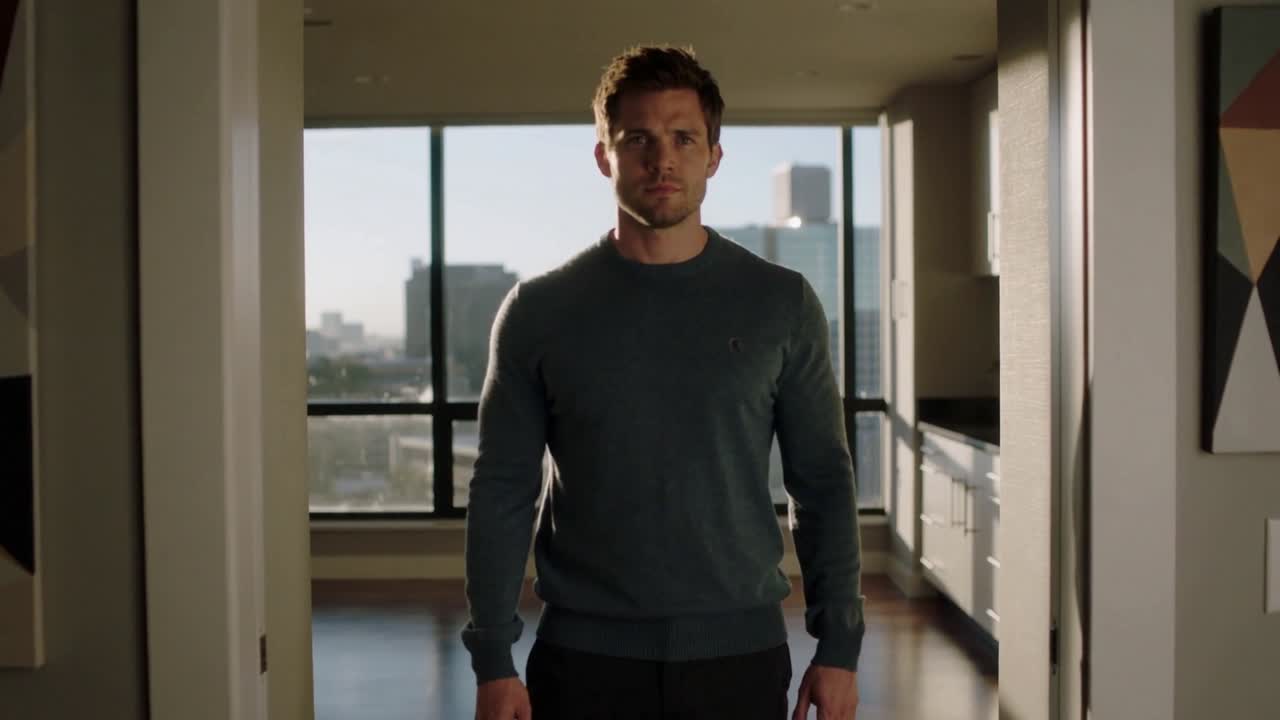 Uomo in maglione girocollo color verde acqua che si avvicina alla telecamera sulla soglia di un appartamento, con una finestra che mostra lo skyline.