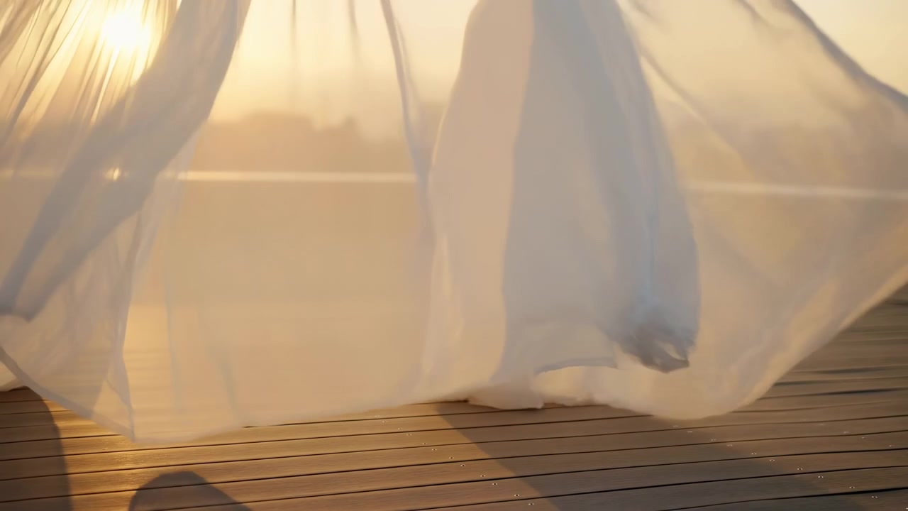 A translucent white fabric billows over a wooden deck in warm sunset light, with a softly blurred city skyline in the background and a gentle sense of motion
