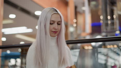 fashion aware mother walks to bench in mall, opens bag to retrieve book, flips pages and reads with serene expression against blurred retail background, capturing stylish leisure moment