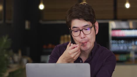 hombre asiático con una computadora portátil tomando un bocadillo y hablando en un teléfono inteligente mientras está sentado en una cafetería
