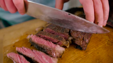 close up view of male hand with a knife slicing the grilled beef steak on cutting board on wooden background