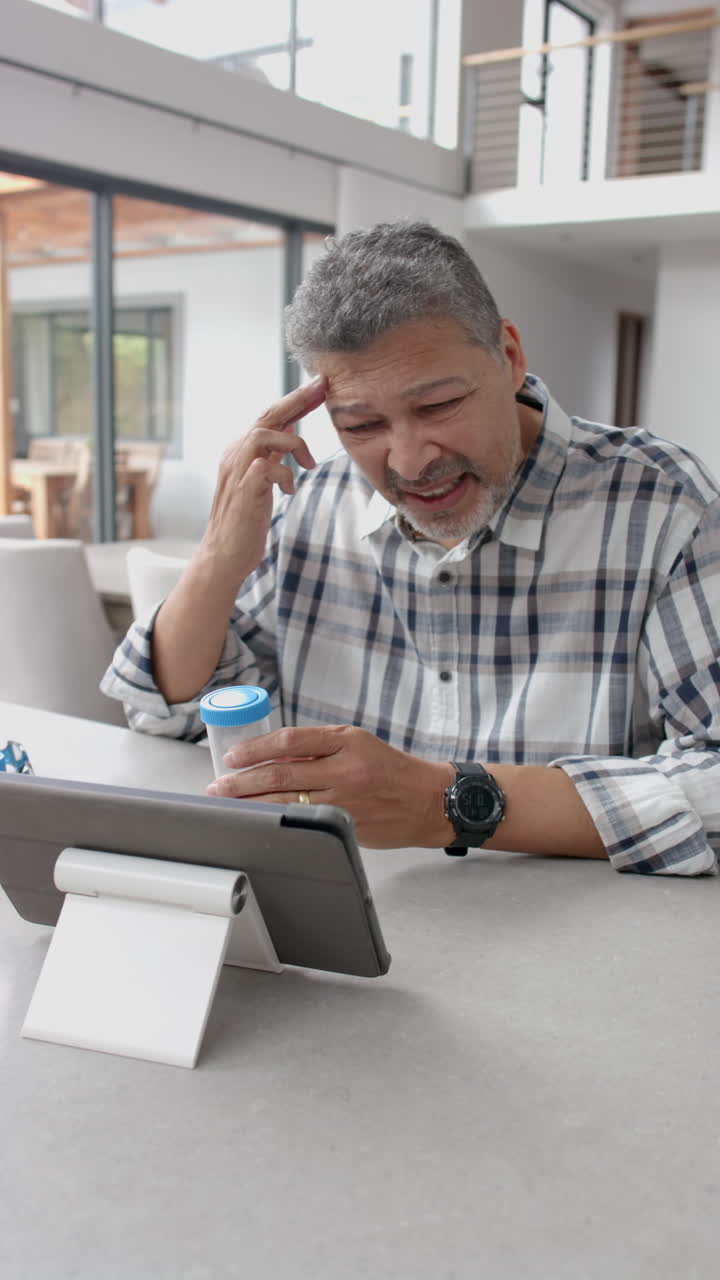 Vertical video of senior biracial man having medical videocall, holding medicine container at home