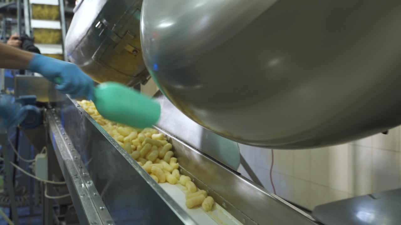Pouring corn sticks of snacks from a metal tank onto an automatic conveyor belt. Hand of a worker with a shovel. Food factory