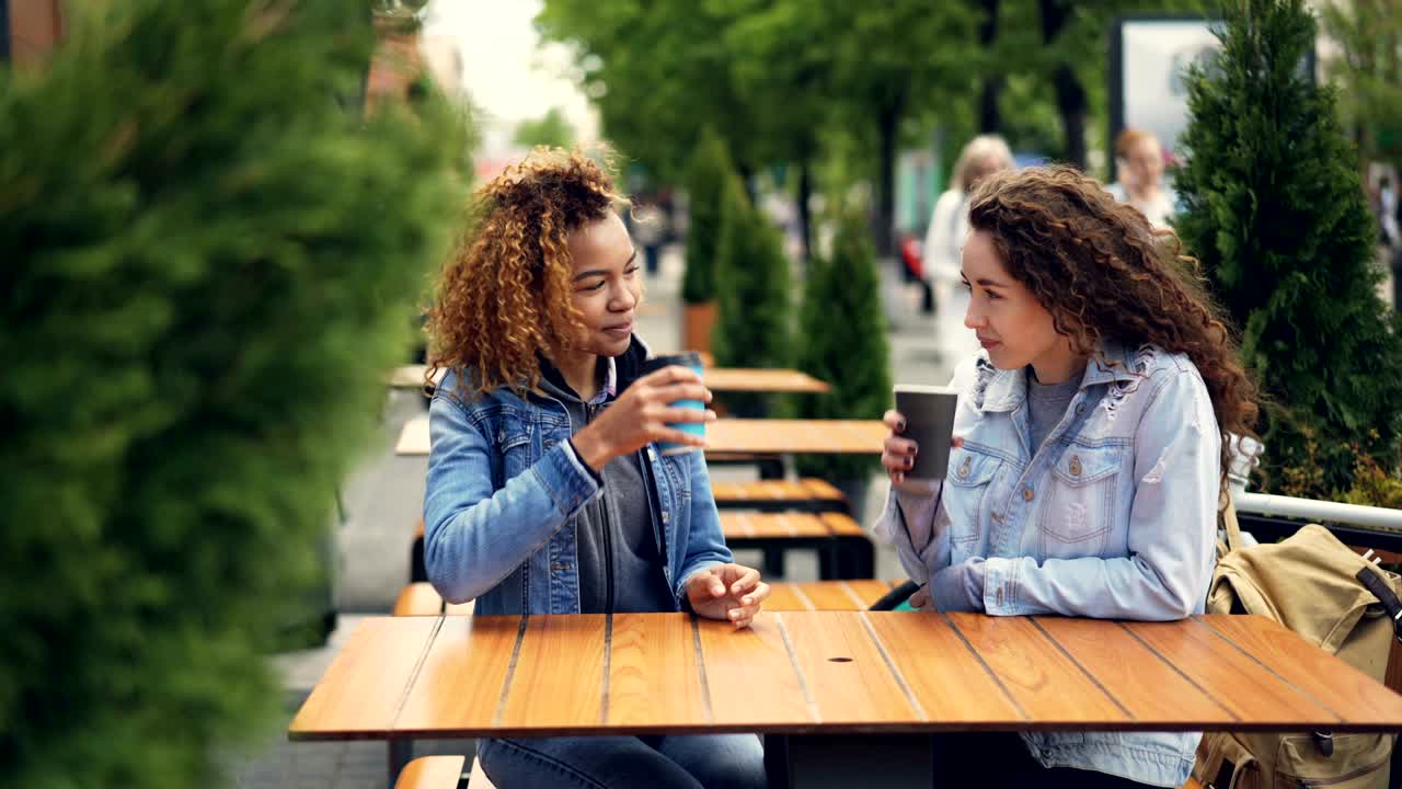 mujeres jóvenes bonitas están brindando y haciendo sonar tazas de café luego bebiendo café y hablando mientras descansan en un café al aire libre en el parque de la ciudad. la gente está caminando en el fondo.