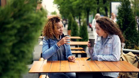 pretty young women are toasting and clinking coffee cups then drinking coffee and talking while resting in outdoor cafe in city park. people are walking in background.