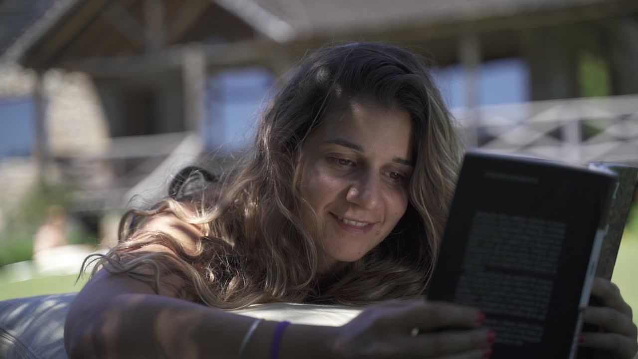 joven mujer latina sonriendo leyendo un libro al aire libre en el césped con vegetación y fondo de casa a cámara lenta 60 fps