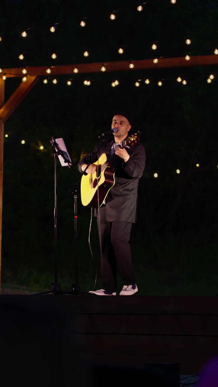 Musiker synger og spiller akustisk guitar på udendørs scene med lyskæder om natten