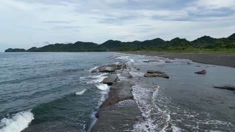 aerial view of crashing waves hitting coastal rocks at idyllic beach with mountains and cloudy skies in the background