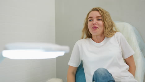 client with long blond hair sits comfortably on leather-covered chair and smiles calmly beside bright white salon lamp in clean, minimalistic interior, preparing for professional beauty treatment