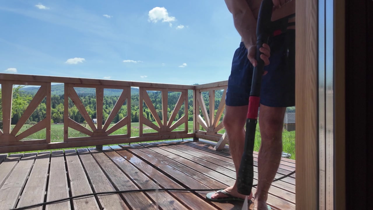 Shirtless man wearing blue shorts washes a wooden terrace with a pressure washer at a log cabin under a bright blue summer sky.