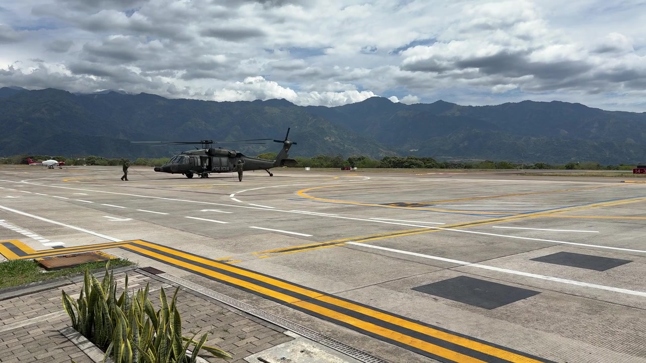 Un elicottero nero della Polizia Nazionale Colombiana rulla sul piazzale, seguito da un camion dei vigili del fuoco dell'aeroporto di colore verde brillante che percorre la pista dell'aeroporto Perales a Ibagué, in Colombia, con lo sfondo in lontananza