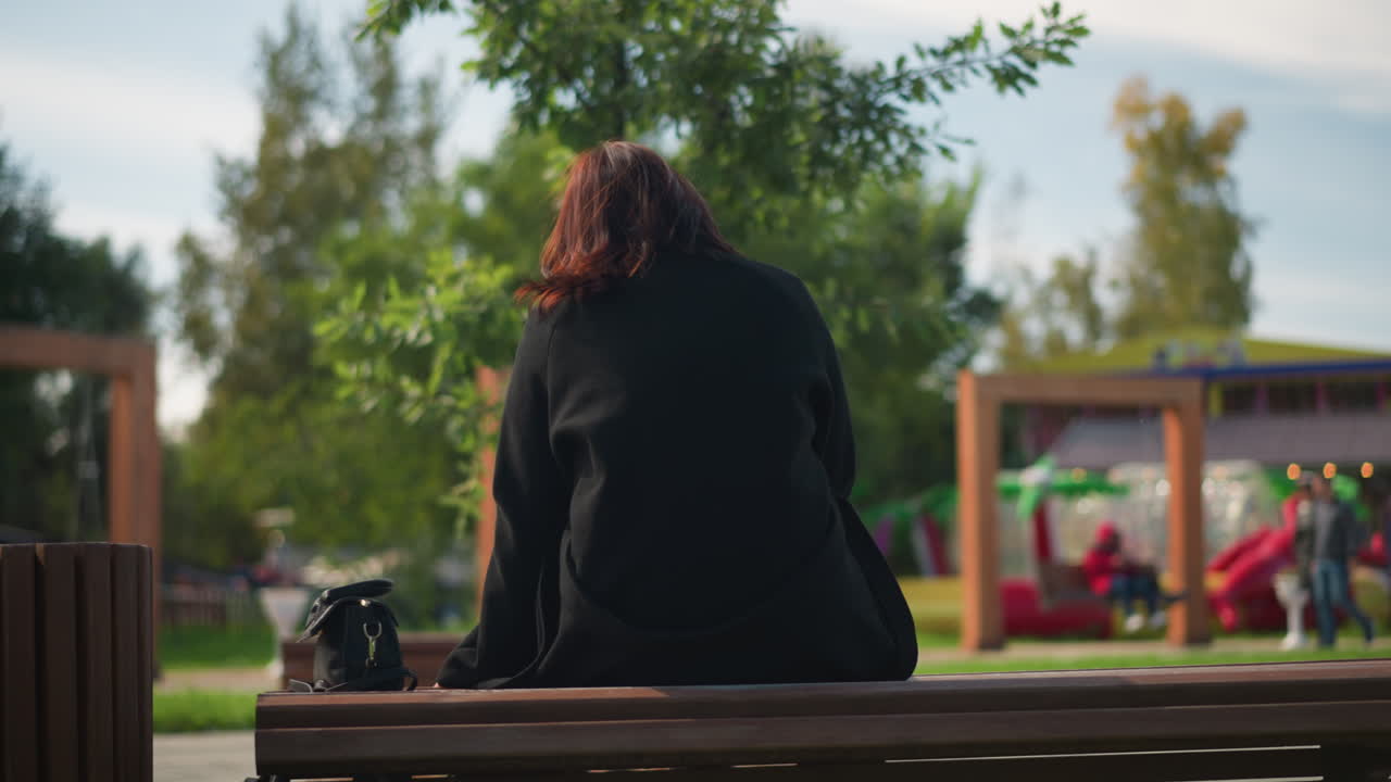 도시 공원의 공원 벤치에 앉아있는 검은 코트와 부츠를 입은 여성의 뒷면, 녹색 나무로 둘러싸여, 놀이터와 함께 흐릿한 배경과 사람들이 돌아다니고 있습니다.