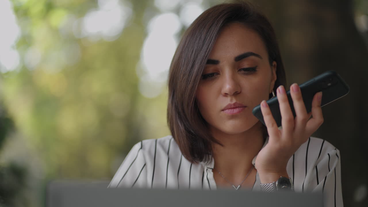 mujer de negocios hispana multitarea que usa la función de reconocimiento de voz en un teléfono inteligente de pie cerca del escritorio y escribiendo en la computadora portátil. empresaria árabe que envía un mensaje vocal