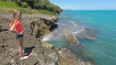 young girl sprayed by mist from natural blowhole on rocky coast of isle of pines