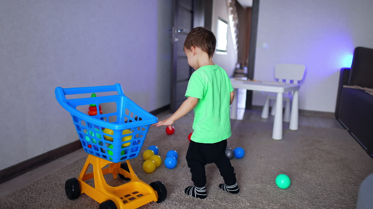 Toddler boy collecting colorful balls into a toy shopping cart. Well-brought child collecting toys in his room.