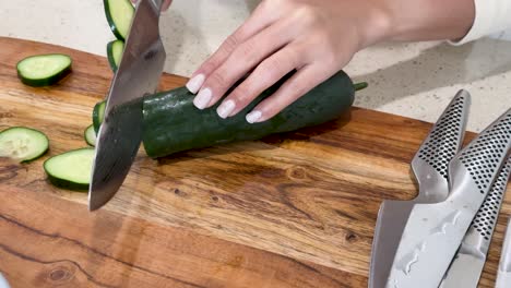 a person with manicured nails slices a cucumber into thin rounds on a wooden cutting board in a bright kitchen, using a large chef’s knife