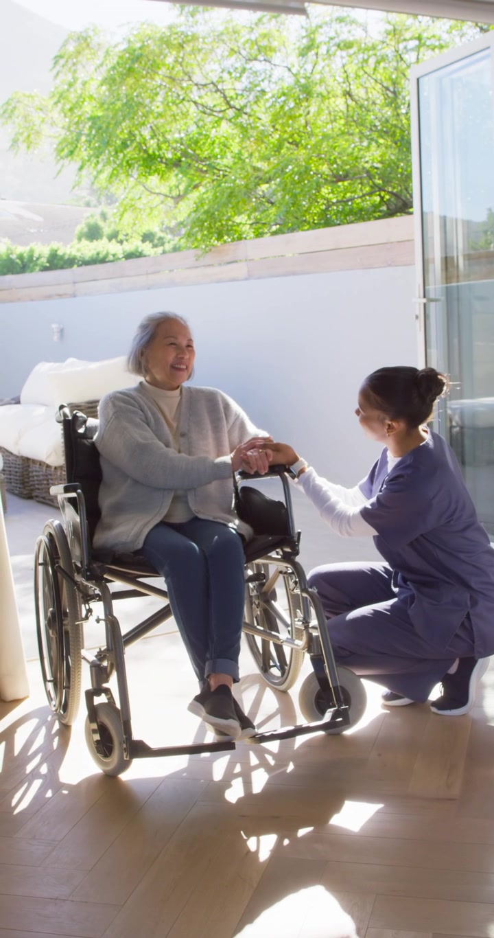 Video verticale: Un assistente si inginocchia accanto a una donna anziana in sedia a rotelle, le tiene le mani per aprire la porta e la conforta.